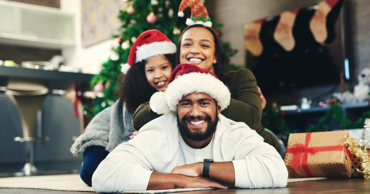 Família de três pessoas usando chapéus de Papai Noel e sorrindo, transmitindo alegria e espírito natalino.