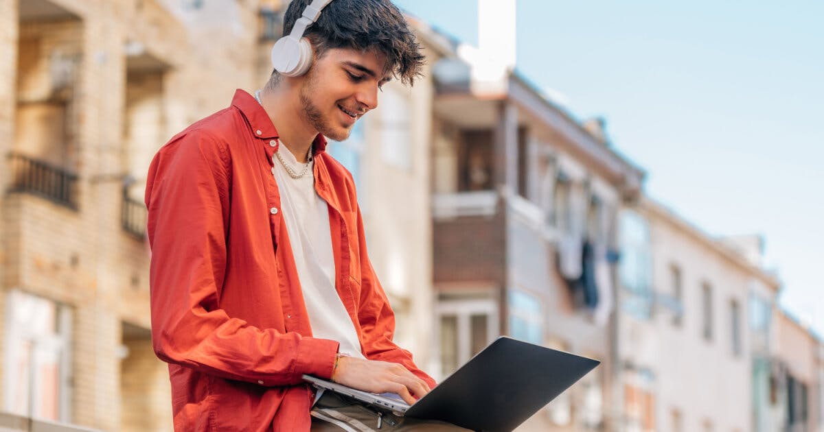 Jovem com fones de ouvido e laptop, concentrado em uma rua movimentada, imerso em sua atividade digital.