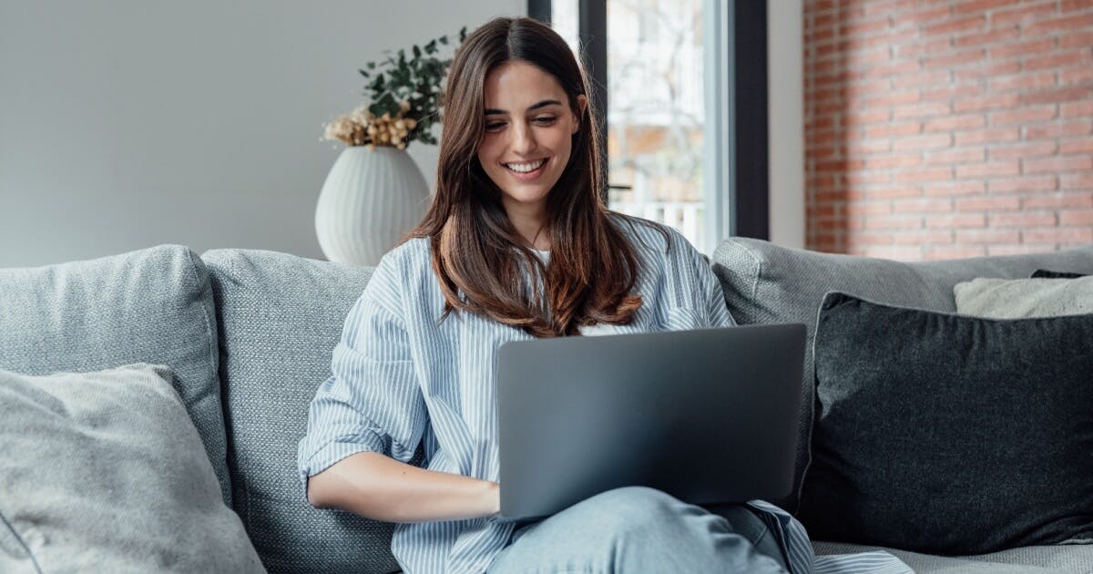 Uma mulher sentada em um sofá, utilizando um laptop, em um ambiente aconchegante e moderno.