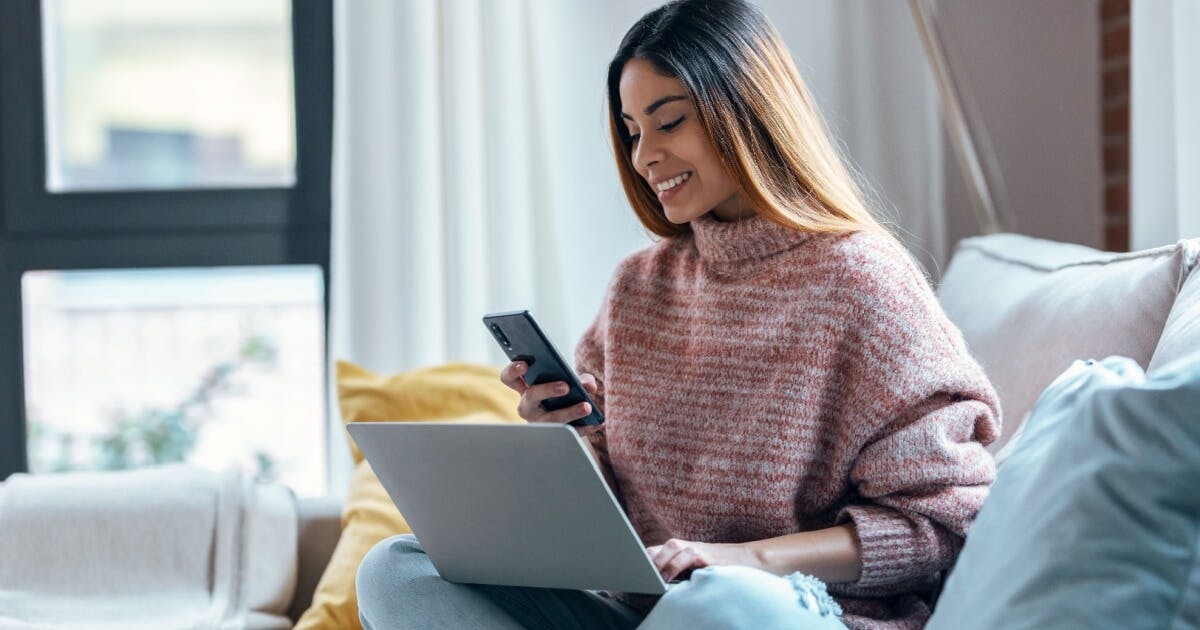 Uma mulher sentada em um sofá, utilizando um laptop e segurando um celular.