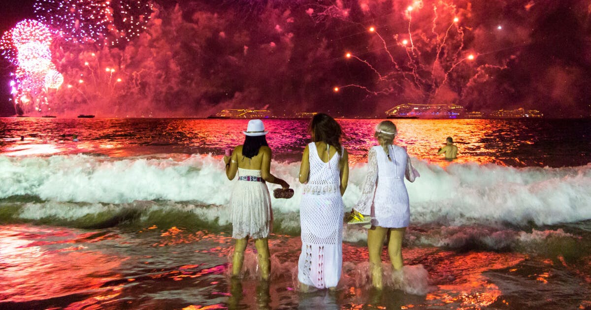 Três mulheres em vestidos brancos na praia, com fogos de artifício iluminando o céu ao fundo.