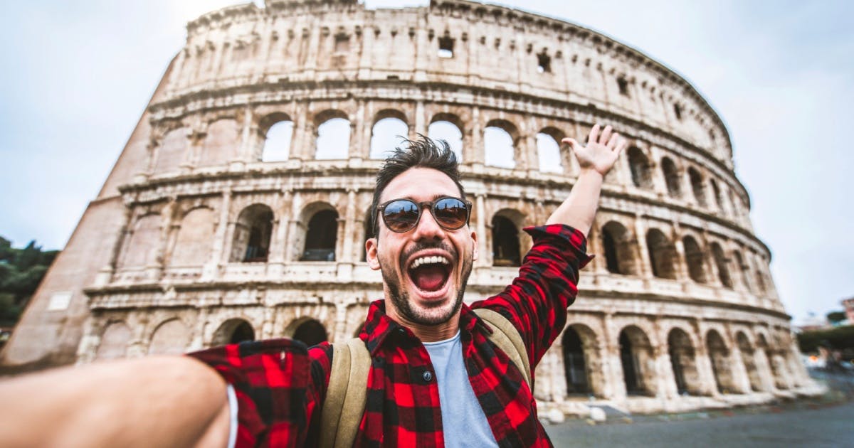 Homem sorrindo e de braços abertos em uma selfie. O coliseu atrás dele contempla a paisagem.