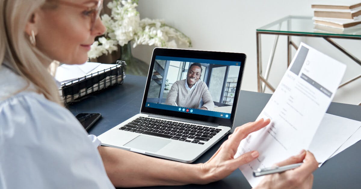 Uma mulher sentada à mesa com um laptop, enquanto um homem aparece na tela do dispositivo.