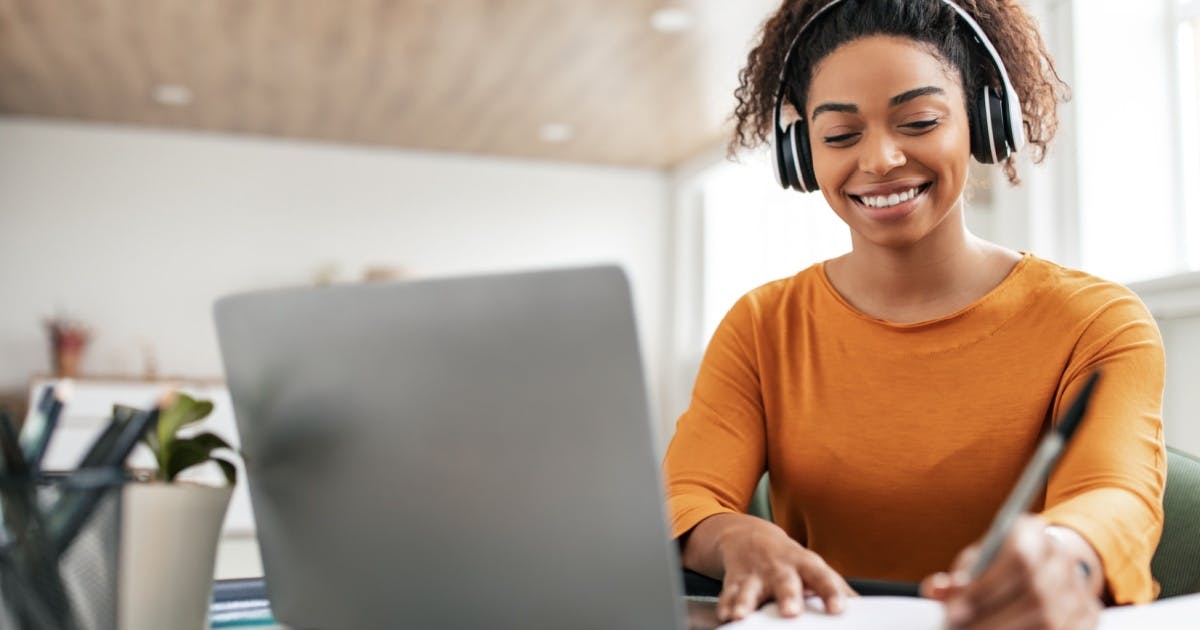 Mulher com uma caneta na mão, fone de ouvido e um notebook aberto na mesa, sorrindo e escrevendo. 