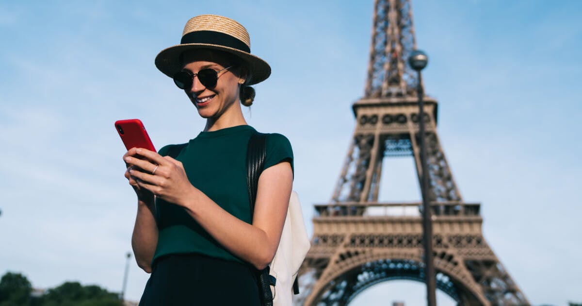 Uma mulher com chapéu e óculos de sol usa o celular em frente à Torre Eiffel.