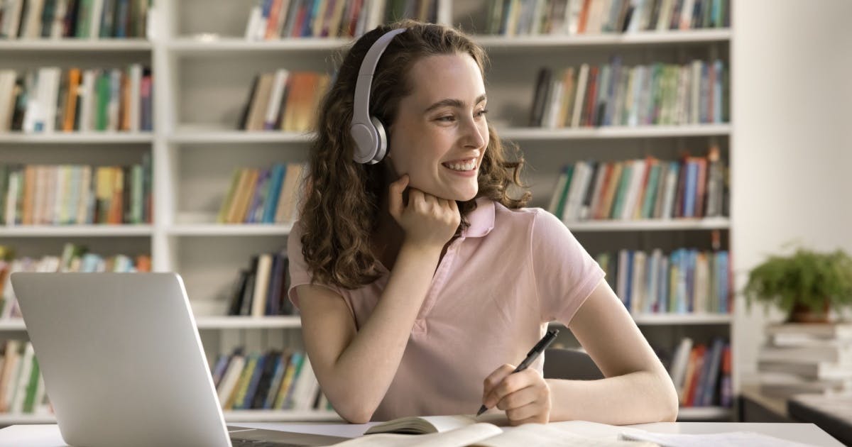 Mulher sorrindo, com fone de ouvido, caneta na mão e um notebook em cima da mesa.