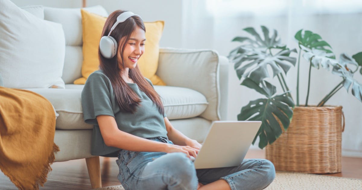 Mulher sentada no chão com um notebook no colo, fone de ouvidos e sorrindo.