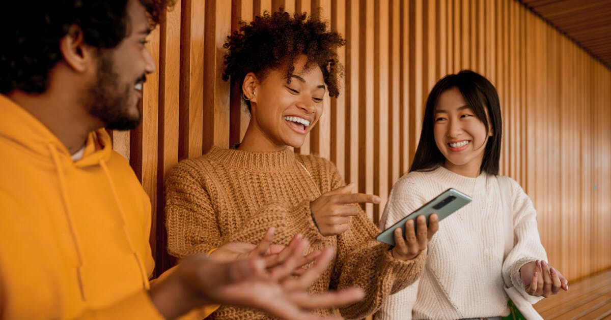 Três jovens sorrindo e rindo enquanto observam um celular, transmitindo alegria e descontração em um momento compartilhado.