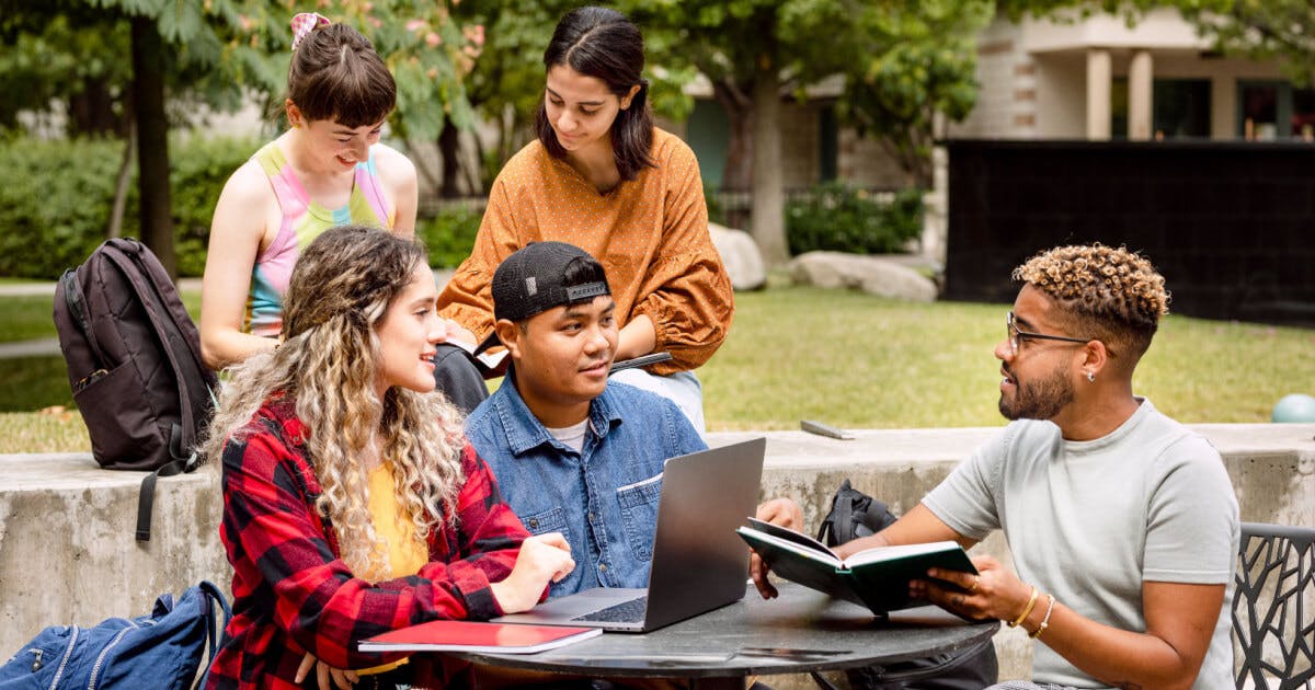 Estudantes concentrados em uma mesa ao ar livre, revisando materiais e trocando ideias em um ambiente agradável.