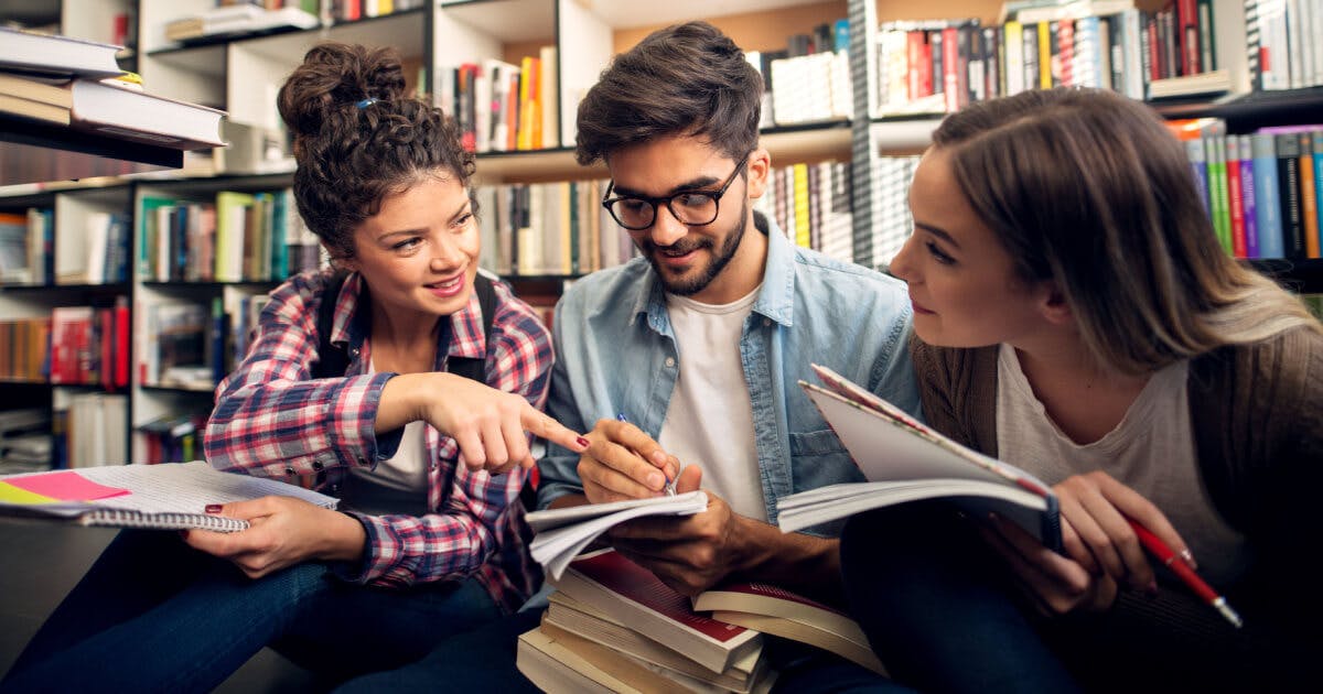 Três jovens sentados em uma biblioteca, cercados por estantes repletas de livros.