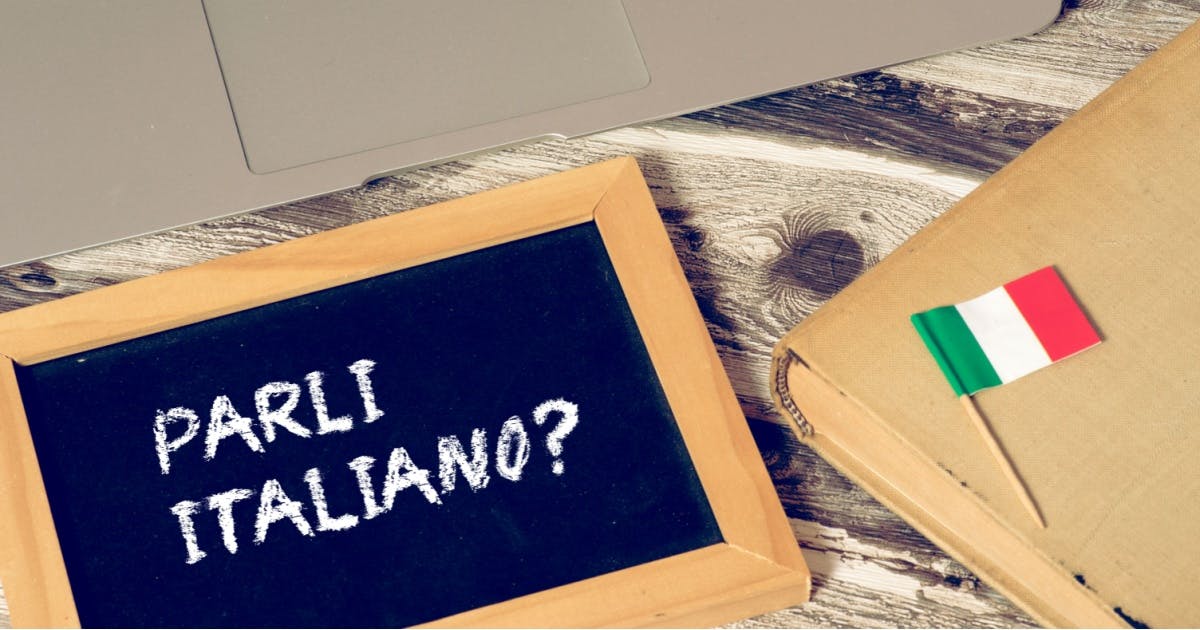 Pergunta em italiano: "Você fala italiano?" em uma pequena lousa ao lado de um notebook para um curso de italiano online.