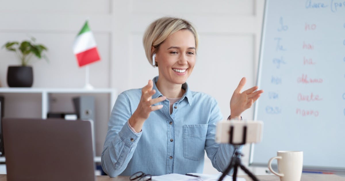 Mulher dando aula em um curso de italiano online, sentada em uma mesa com um laptop e uma câmera, equipamentos de trabalho.