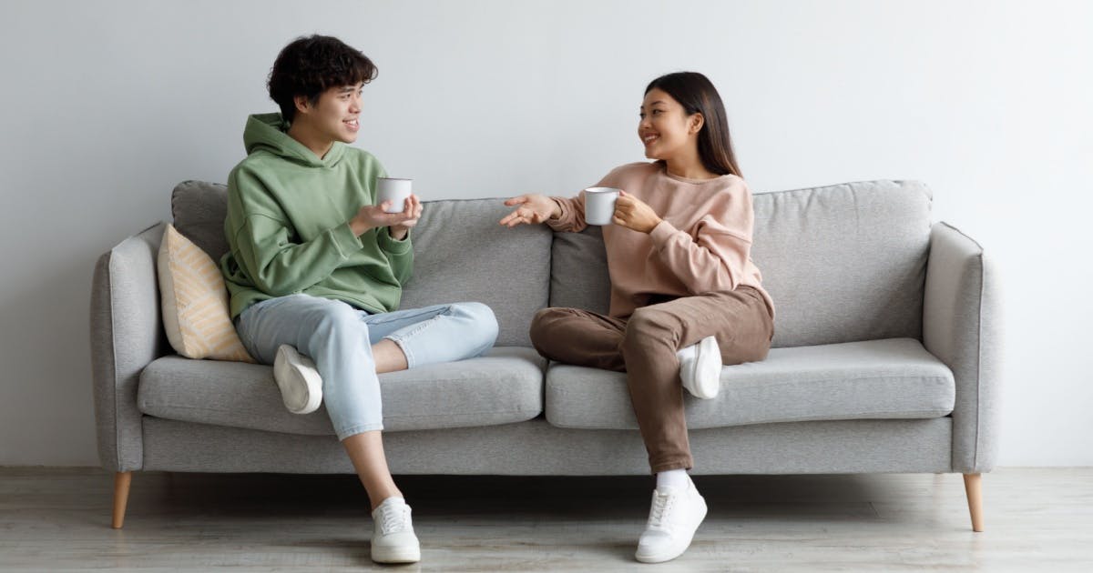 Um homem e uma mulher com café nas mãos, sentados em um sofá e olhando sorrindo um para o outro.