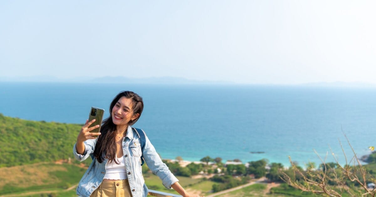 Uma mulher tirando selfie e atrás uma paisagem com floresta e mar
