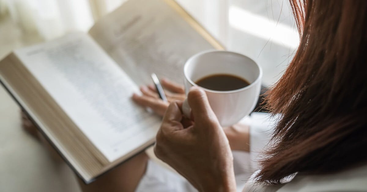 Uma pessoa lendo um livro, com uma xícara de café em uma mão e caneta na outra