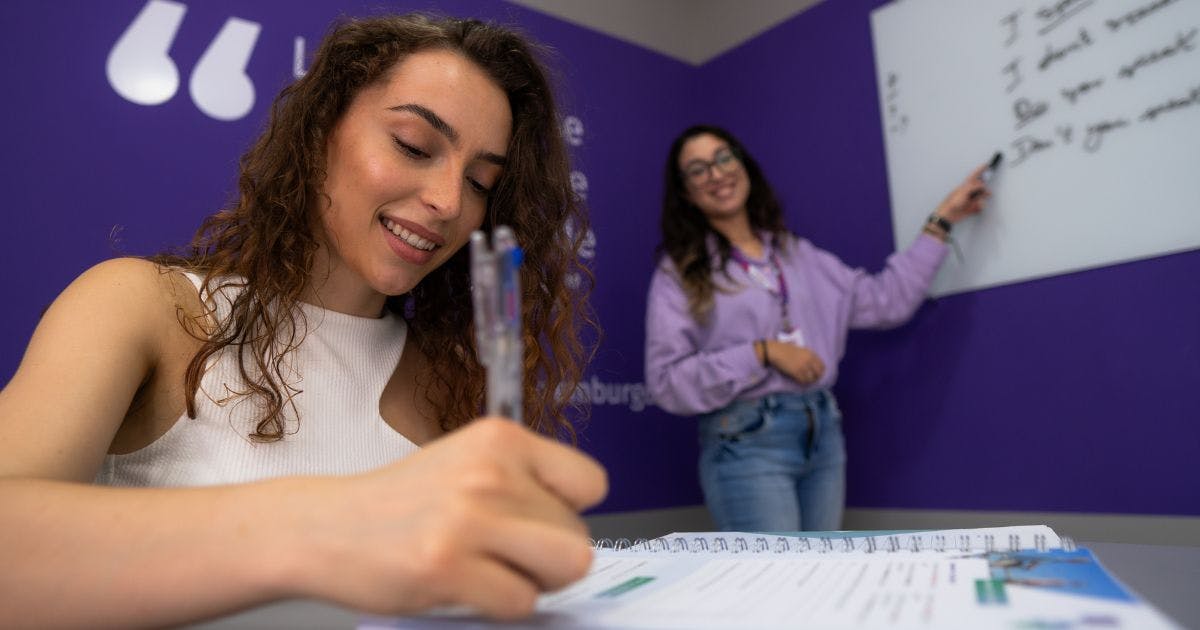 Aluna sorrindo com a caneta na mão e a professora atrás no quadro ensinando