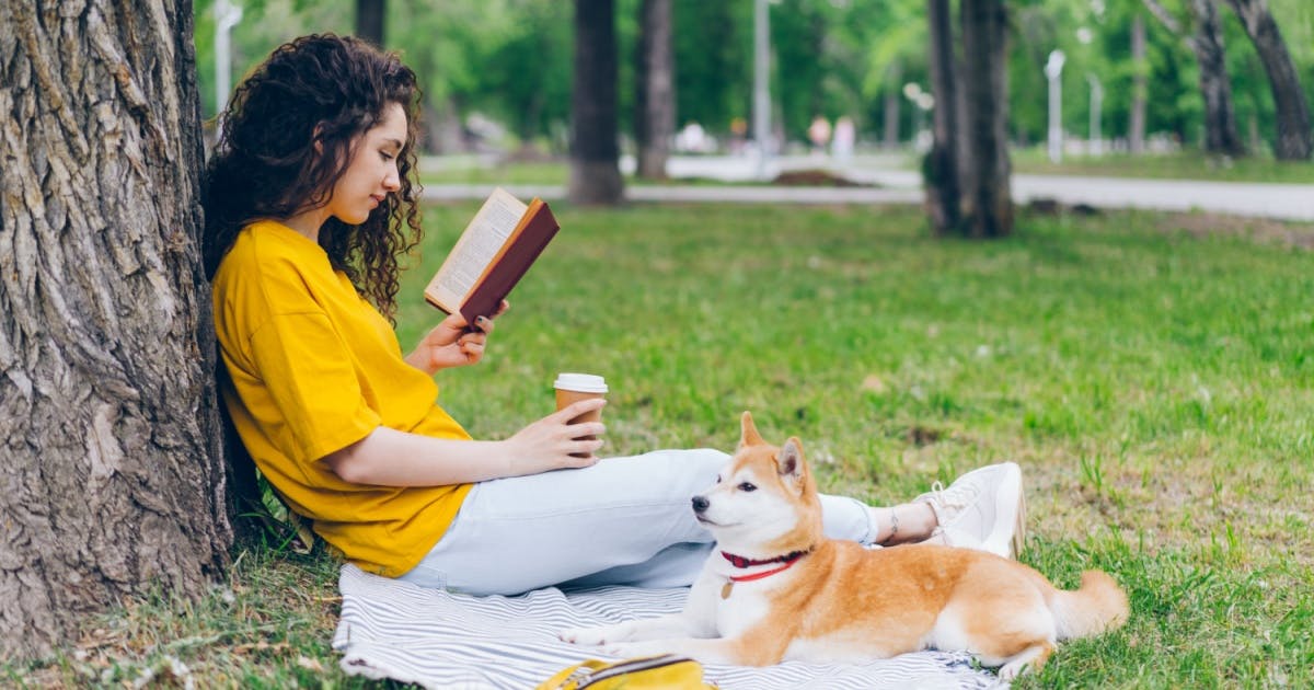 Uma mulher lendo um livro, com um café na mão e um cachorro deitado ao seu lado. 