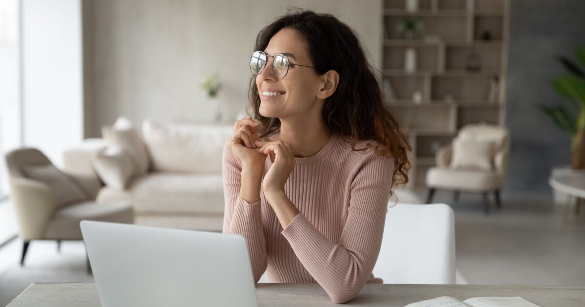 Uma mulher sorrindo e olhando para o horizonte com um notebook em cima de sua mesa.