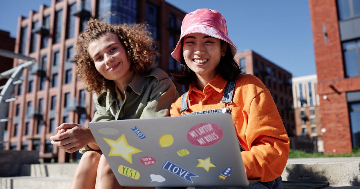 Duas jovens mulheres sentadas em um banco, utilizando um laptop, em um ambiente ao ar livre.