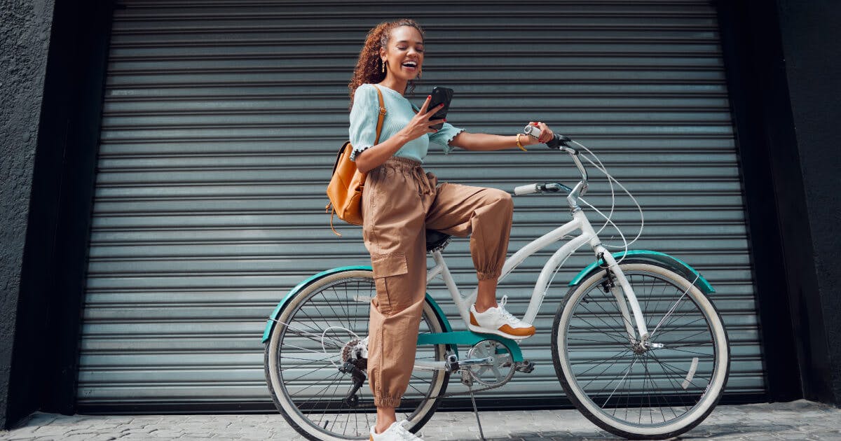 Mulher em uma bicicleta, segurando um celular, enquanto pedala em um ambiente urbano.