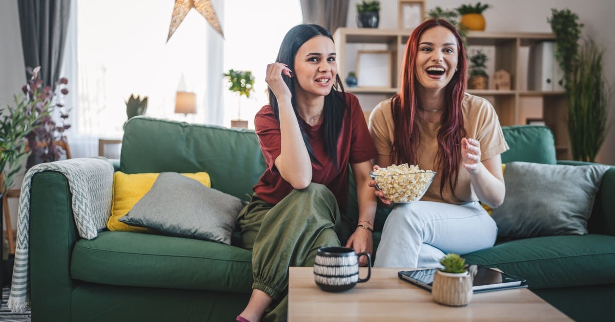 Duas mulheres assistindo TV, com um pote de pipoca na mão e sorrindo.