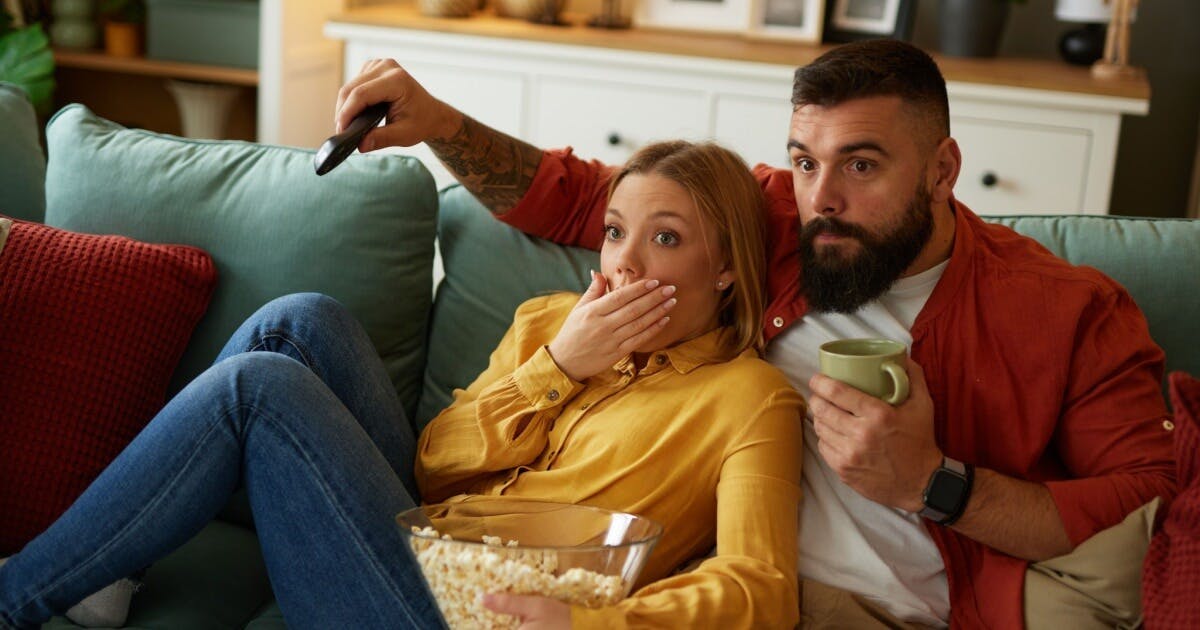 Uma mulher e um homem com expressão de surpresa assistindo a um filme. Ela com uma pipoca na mão e ele com o controle e um café.