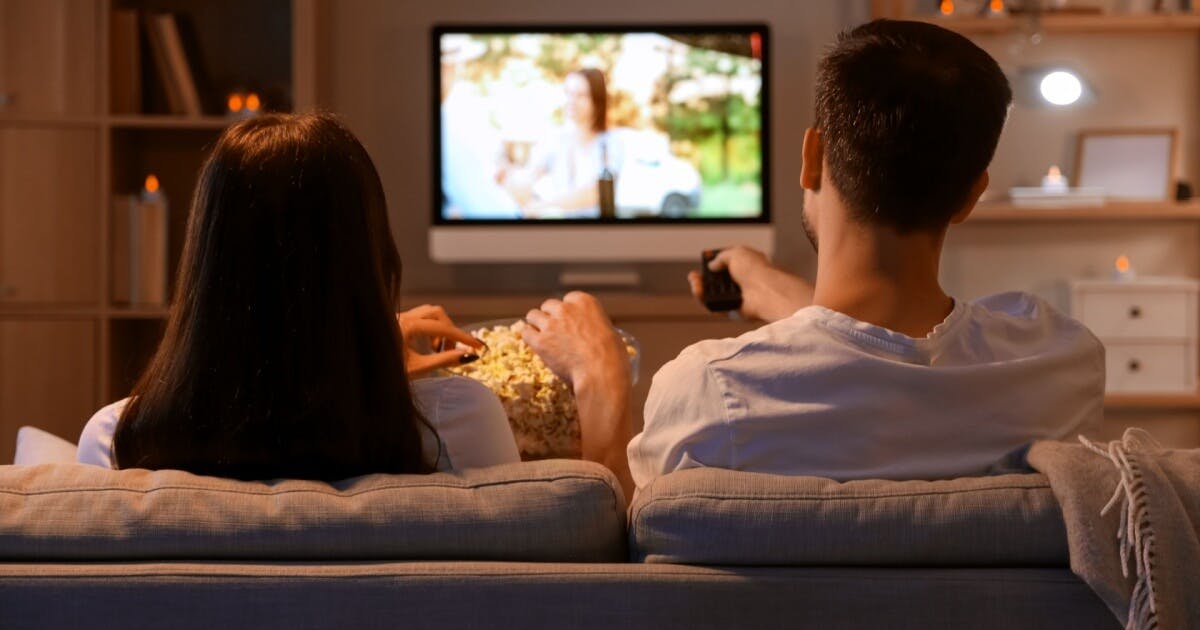 Um casal assistindo filme, ela com um pote de pipoca e ele com o controle na mão.