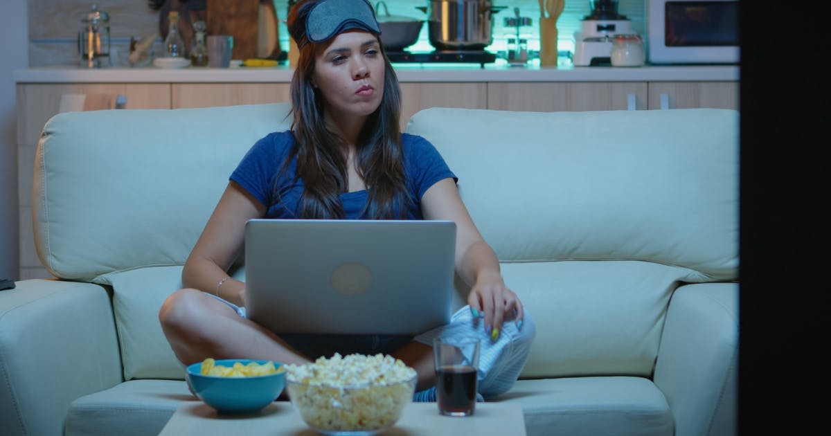 Mulher assistindo filme sentada no sofá com um notebook no colo e um pote de pipoca, um de batatas fritas e um refrigerante ao lado.