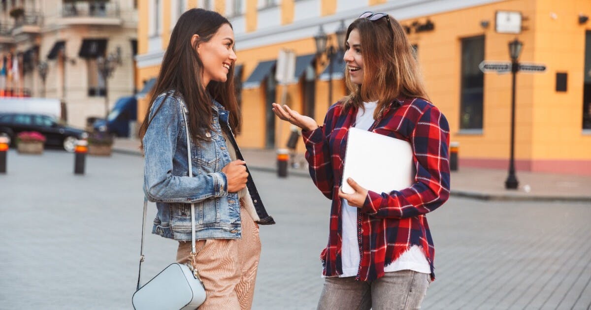 Duas jovens conversando animadamente na rua, com expressões alegres e gestos que demonstram envolvimento na conversa.