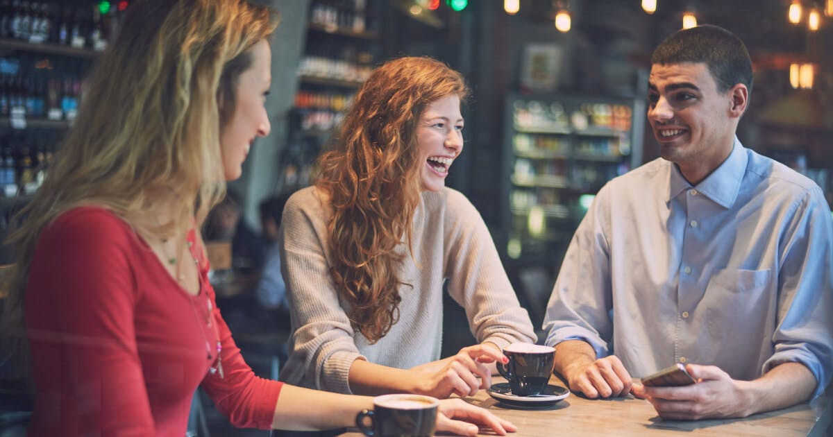 Três pessoas sentadas à mesa em uma cafeteria, conversando e desfrutando de bebidas quentes.