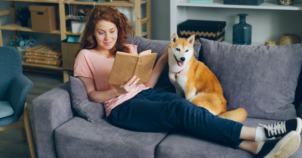 Mulher lendo um livro com seu cachorro no sofá, criando um ambiente aconchegante e relaxante.