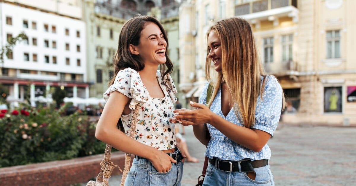 Duas jovens mulheres rindo e conversando animadamente em uma rua movimentada.