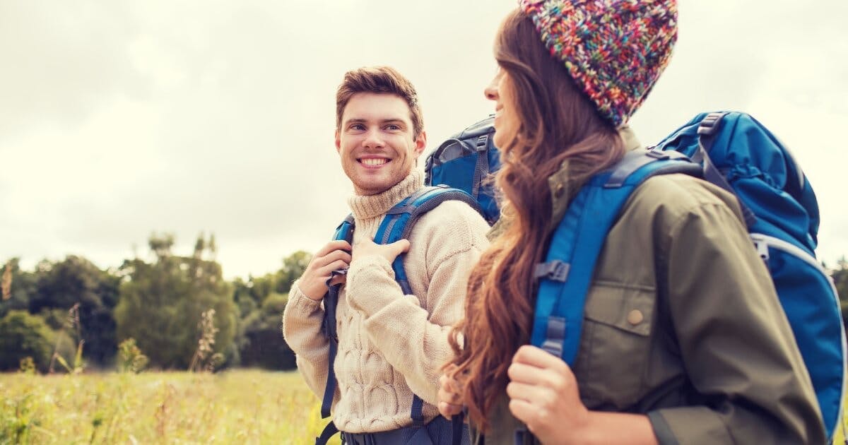 Casal com mochilas caminhando em um campo aberto, cercado por natureza e sob um céu claro.