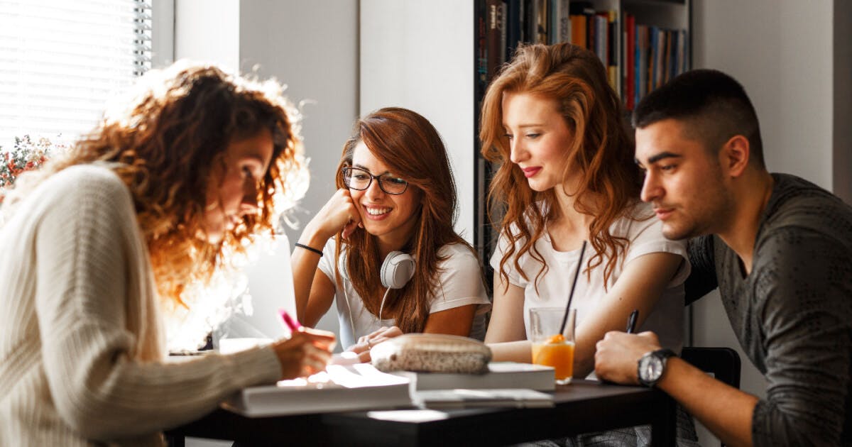 Quatro jovens sentados à mesa com um laptop, envolvidos em uma discussão ou atividade colaborativa.