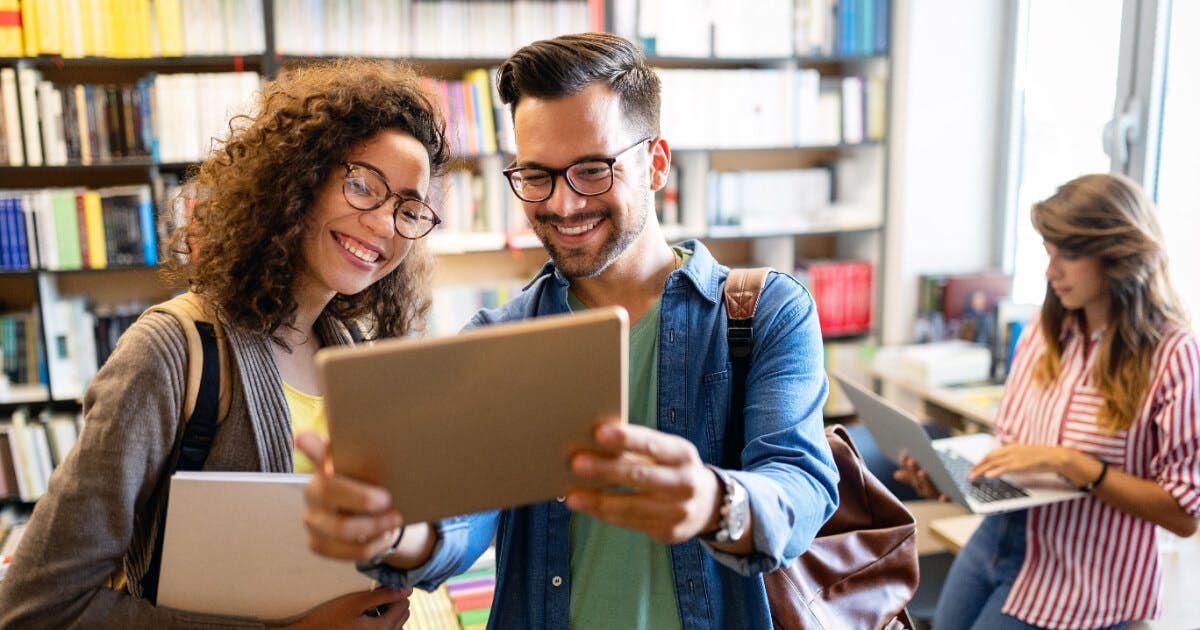 Estudantes utilizando um tablet em uma biblioteca, cercados por prateleiras de livros e um ambiente de estudo tranquilo.