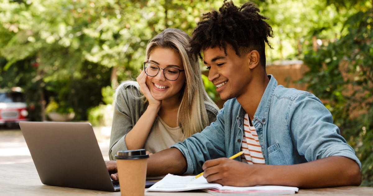 Dois jovens sentados à mesa com um laptop, concentrados em suas atividades.