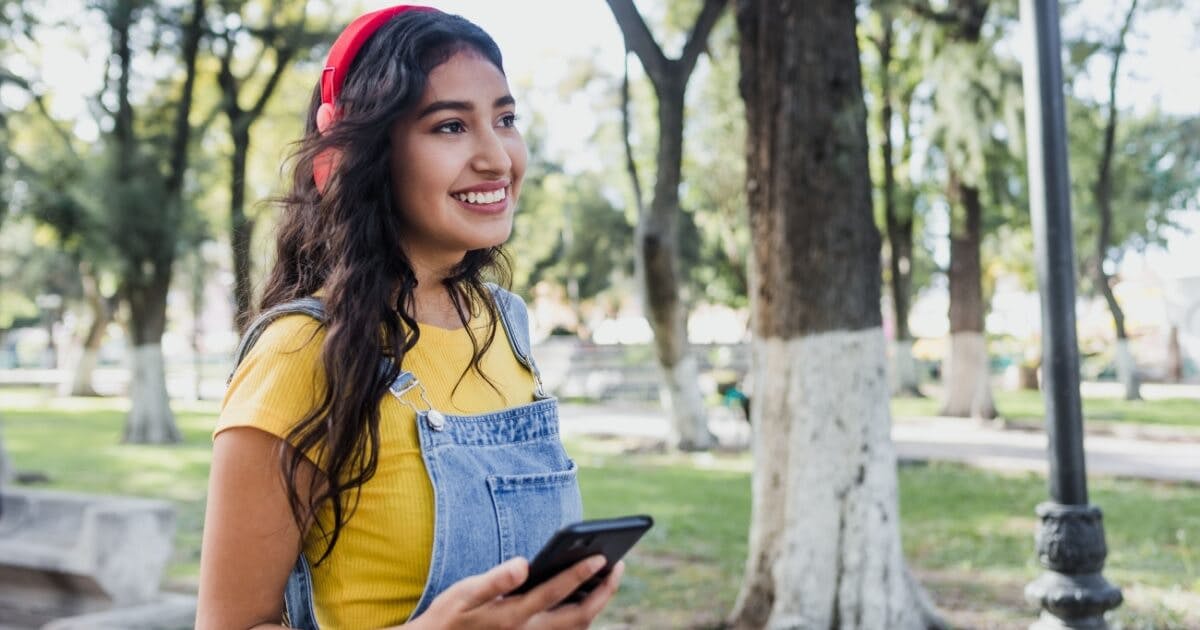 Uma mulher sorrindo, com um celular na mão e um fone de ouvido vermelho.