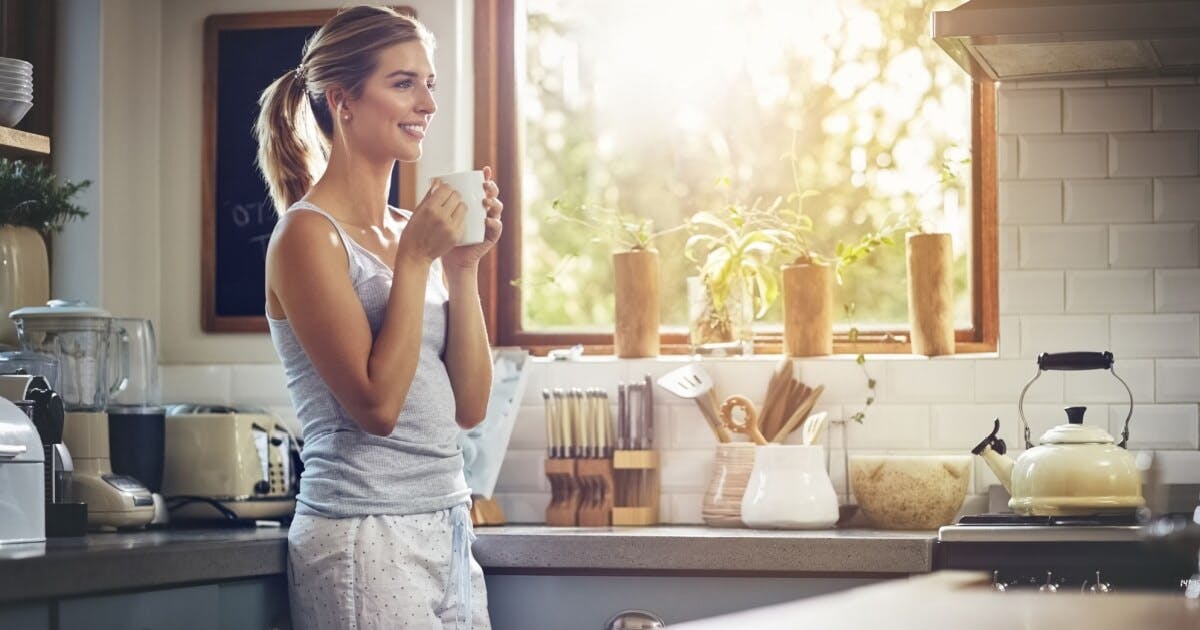 Uma mulher loira segurando uma xícara de café escorada no balcão de uma cozinha.