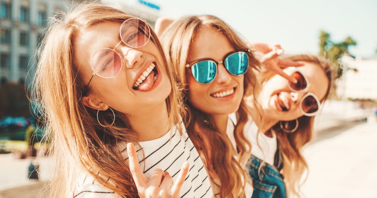 Três jovens mulheres sorrindo e usando óculos de sol, transmitindo alegria e descontração em um ambiente ensolarado.