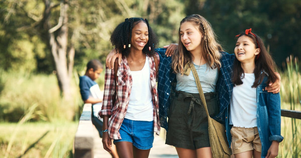 Três meninas jovens em uma ponte, uma delas sorrindo, transmitindo alegria e amizade em um cenário ao ar livre.