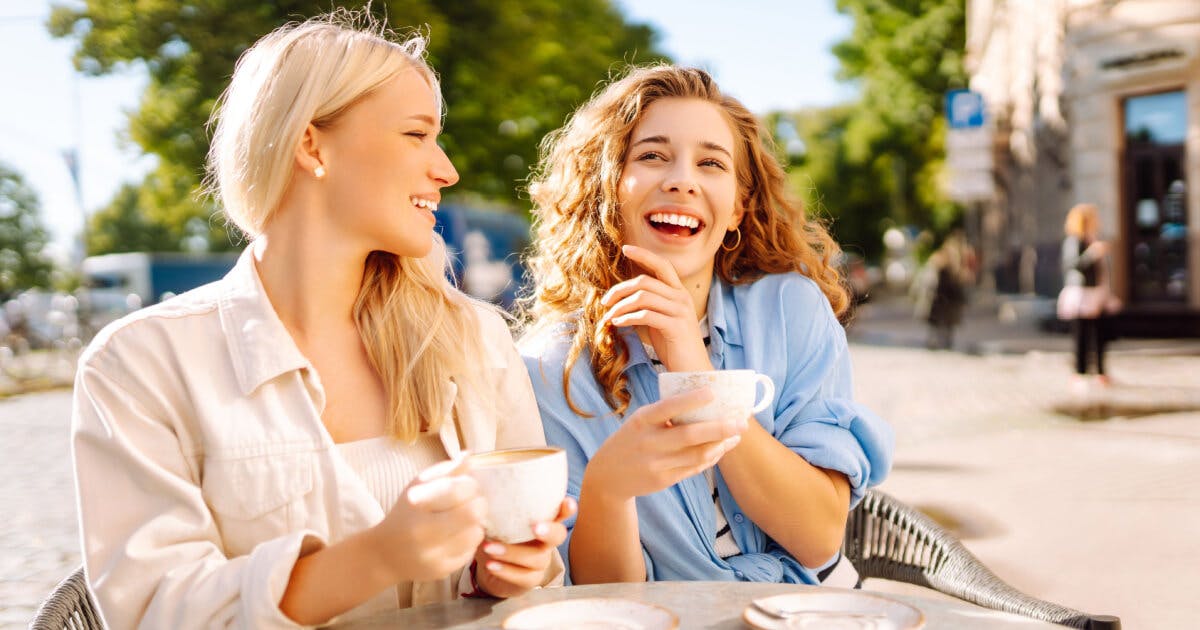 Duas mulheres sentadas em uma mesa ao ar livre, desfrutando de café e conversando animadamente.