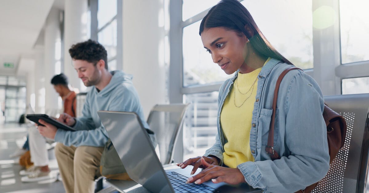 Uma mulher sentada em um aeroporto, utilizando seu laptop, cercada por malas e pessoas ao fundo.