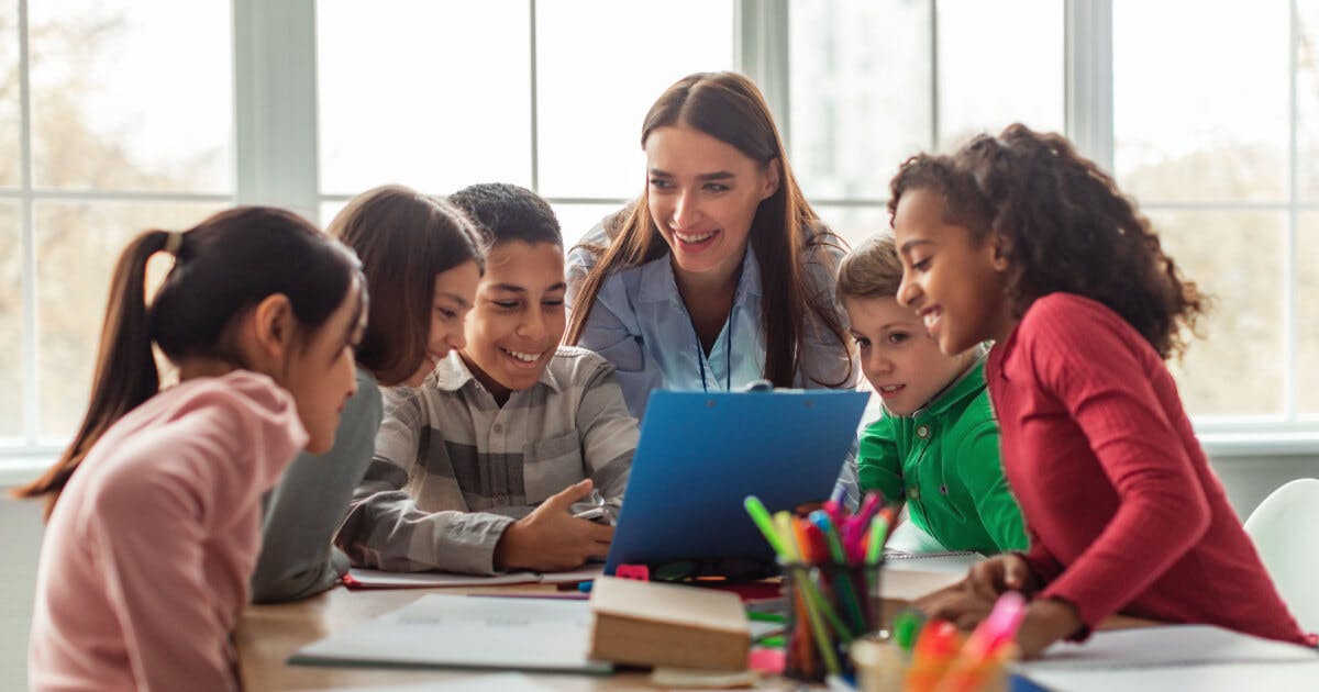 Professor e alunos colaborando em um laptop durante uma atividade de aprendizado em sala de aula.