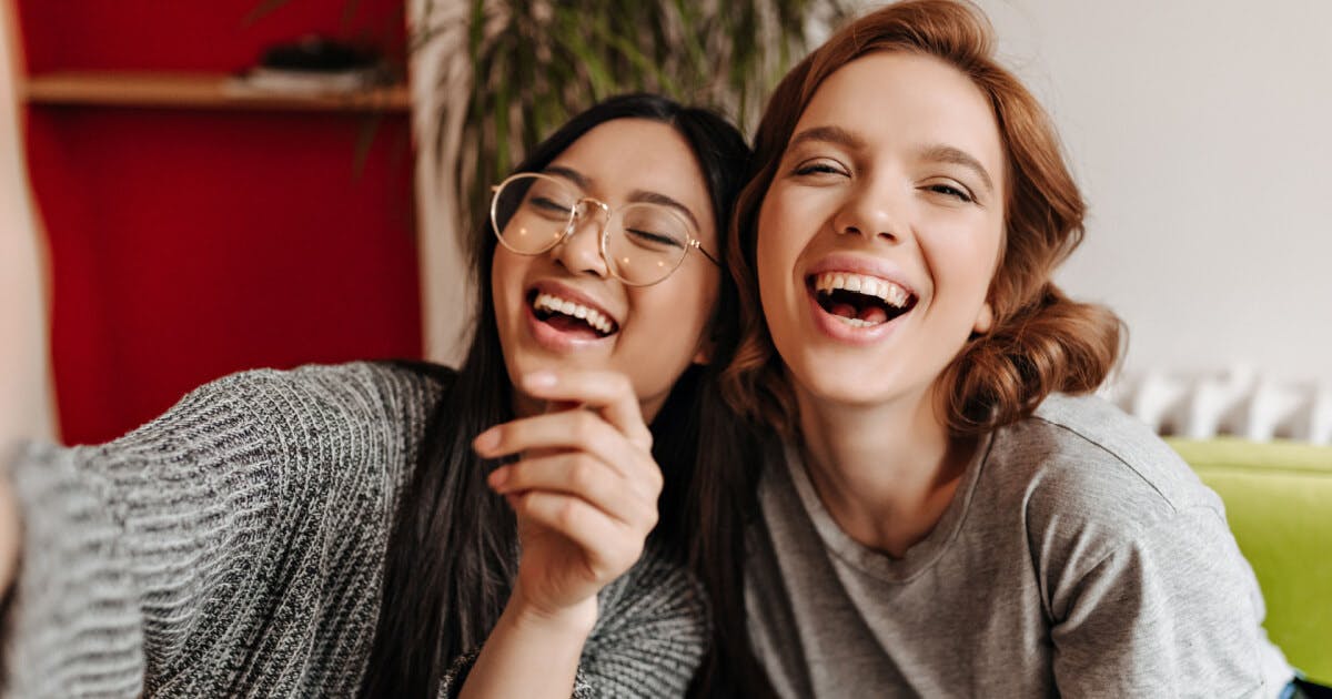 Duas mulheres sorrindo e rindo enquanto estão sentadas em um sofá, transmitindo alegria e descontração.