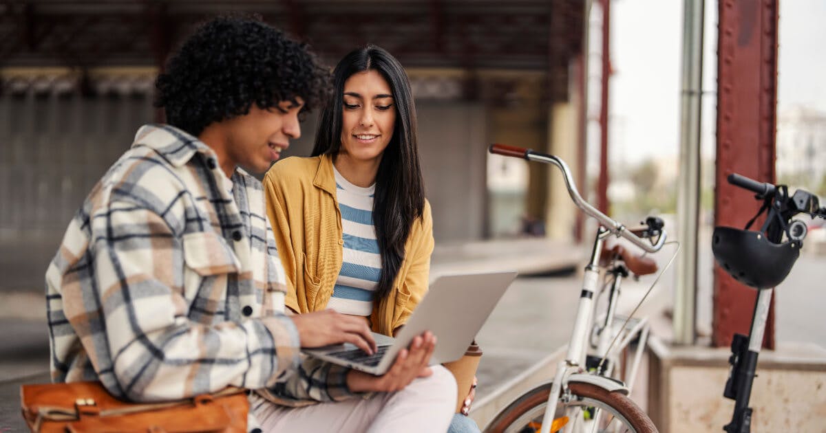 Dois jovens sentados em um banco, utilizando um laptop, em um ambiente ao ar livre.