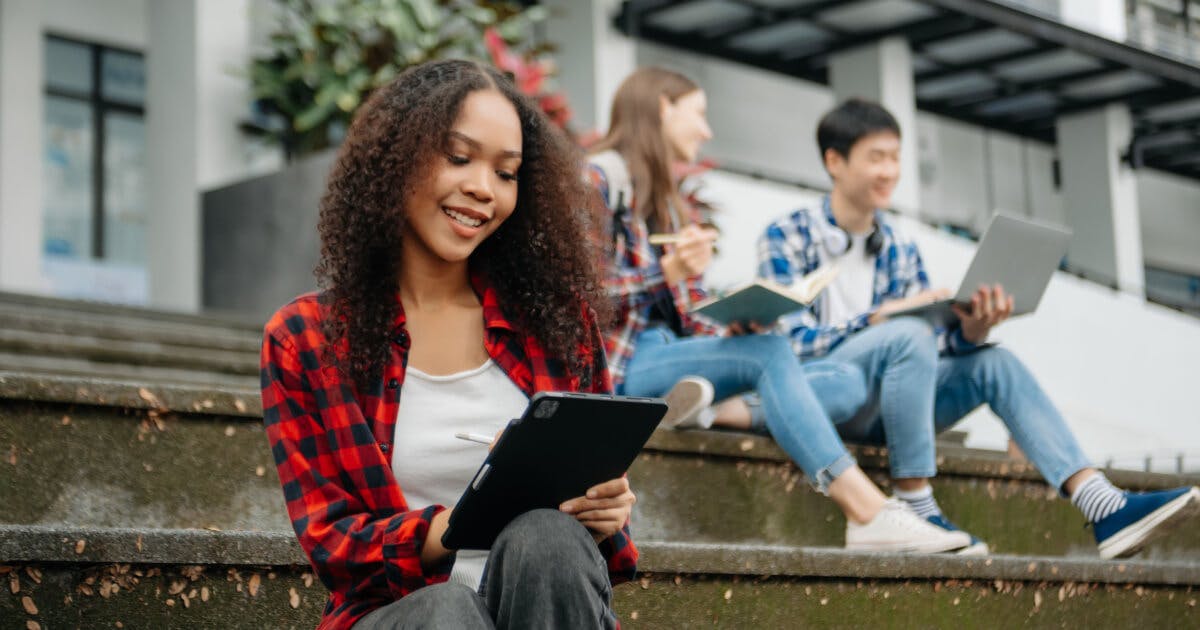 Uma jovem mulher sentada em degraus, utilizando um laptop, com expressão concentrada e ambiente ao ar livre.