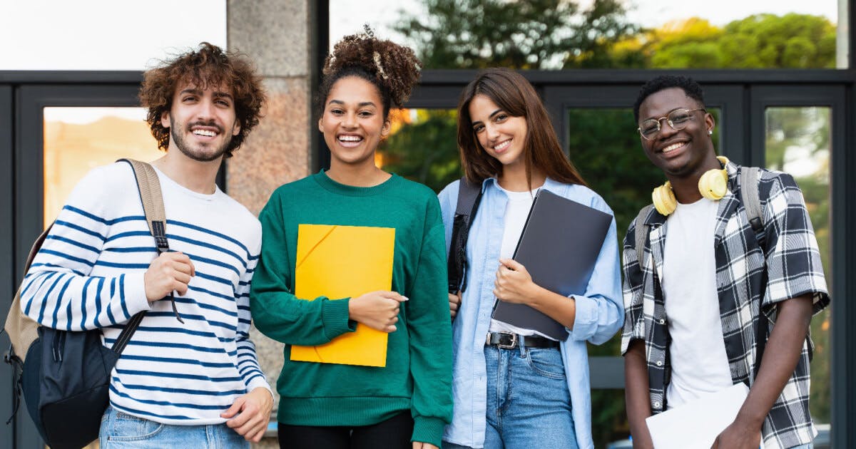 Quatro jovens em pé em frente a um edifício, sorrindo e interagindo entre si, em um ambiente urbano.