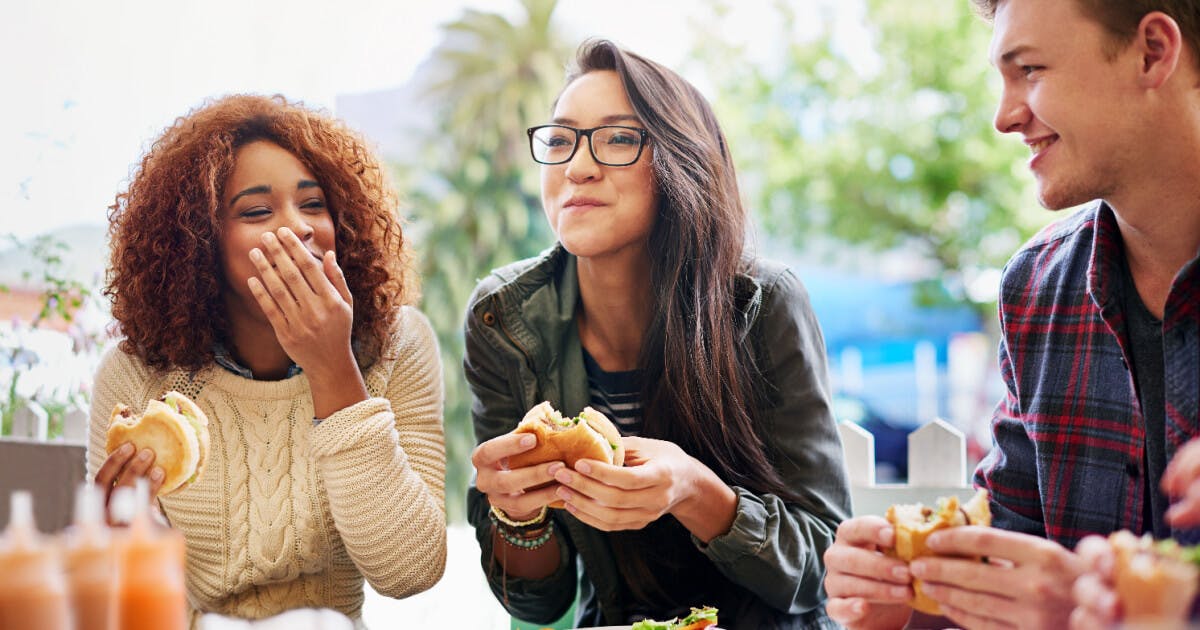 Três jovens saboreando sanduíches em uma mesa, compartilhando momentos de descontração e amizade.