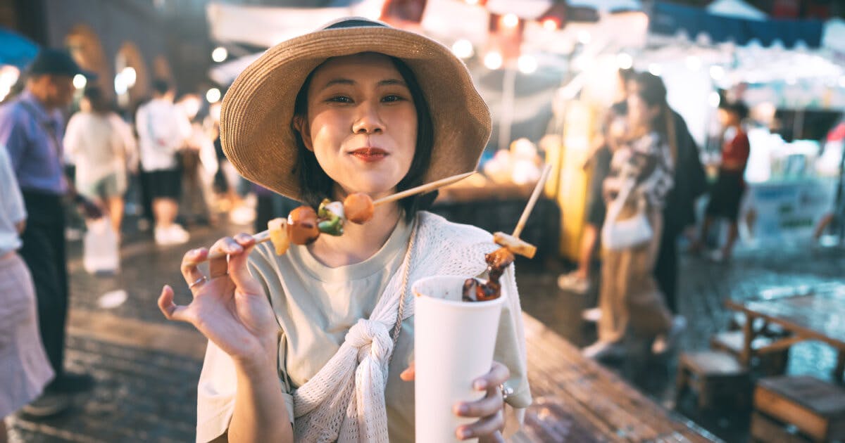 Mulher asiática saboreando comida em um mercado ao ar livre, cercada por barracas coloridas e um ambiente vibrante.