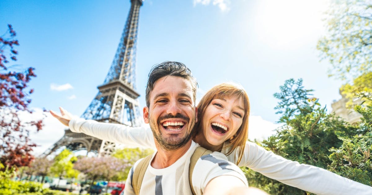 Um homem e uma mulher (de braços abertos) sorrindo e atrás a Torre Eiffel, em Paris, na França.